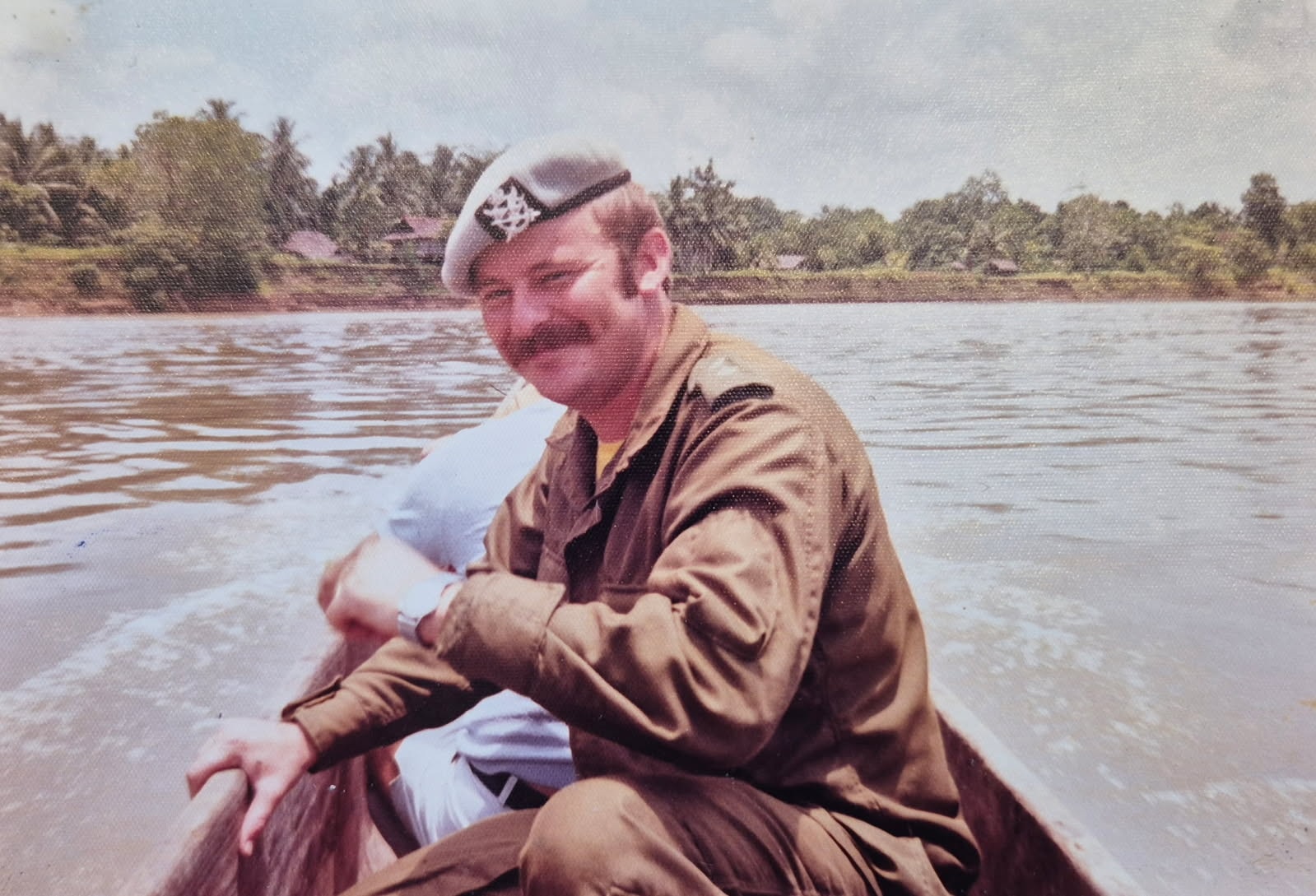 A young man in army uniform and a beret sits in a dugout canoe on a wide brown river, smiling back over his shoulder, a thatched village on the far bank.