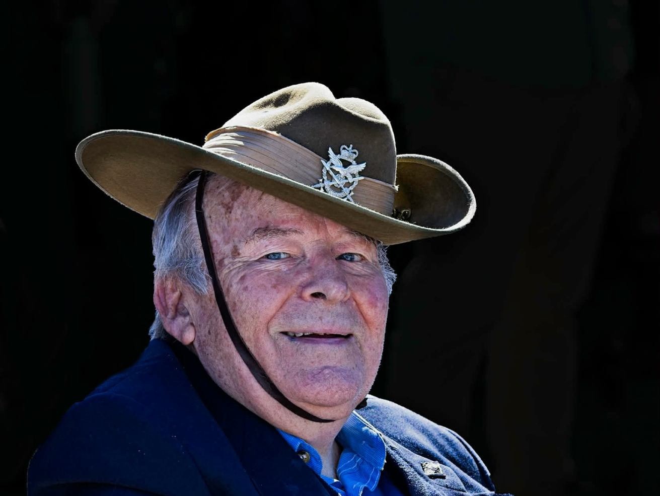 An older man in a slouch hat with a Rising Sun badge, chin strap under his jaw, smiling directly at the camera against a dark background.
