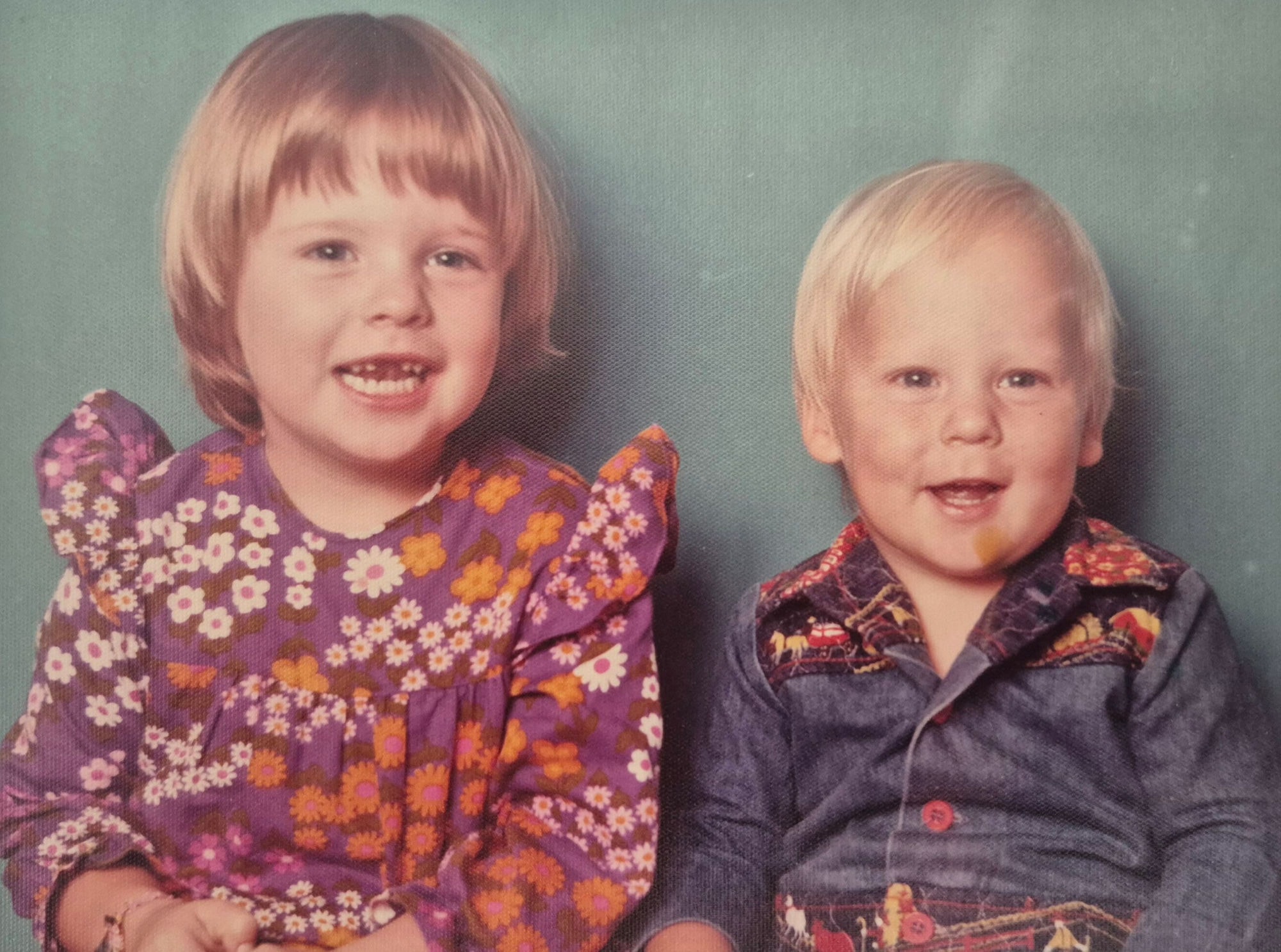 A 1970s studio portrait of two small children, a girl in a purple floral dress beside a younger boy in a denim shirt with a horse-print yoke.