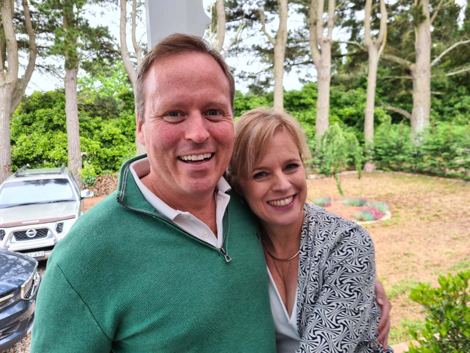 Damien and Liane together outdoors among pine trees, both smiling at the camera.