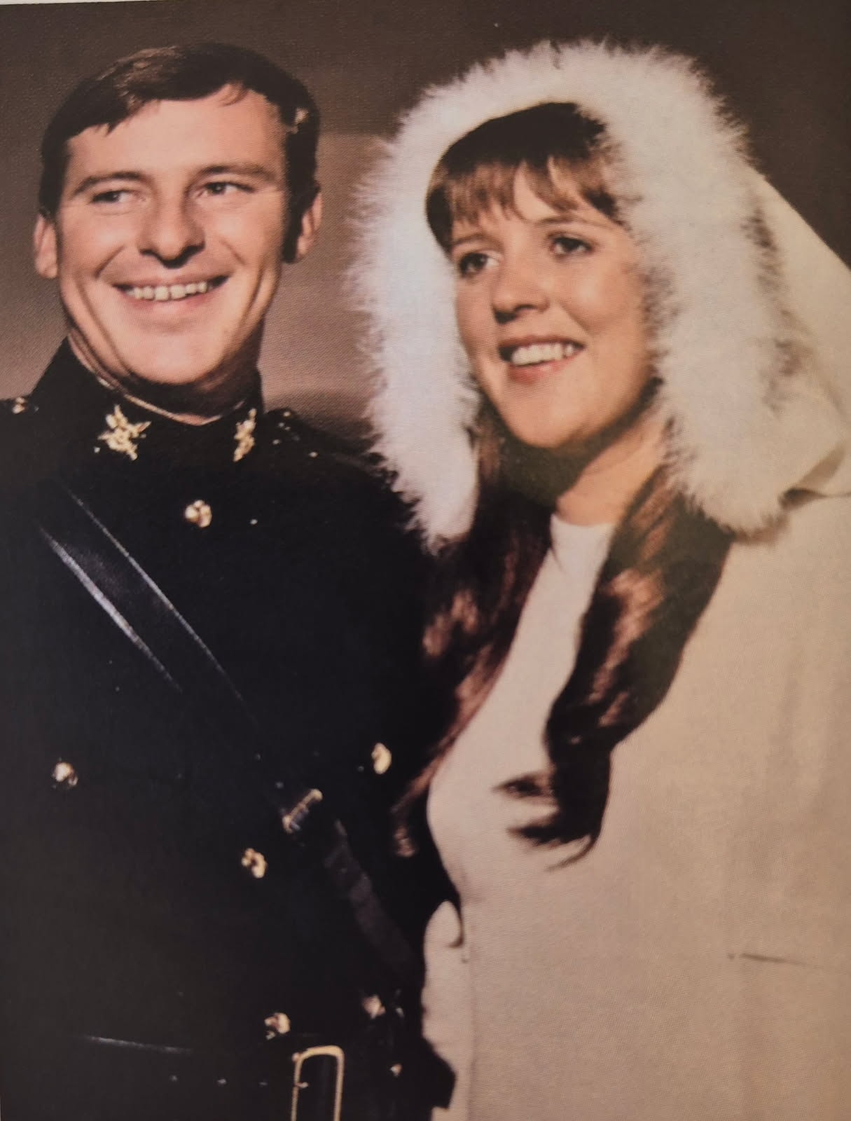 A young man in dark military mess dress with a sash beside a young woman in a cream coat with a white fur-trimmed hood, both laughing.