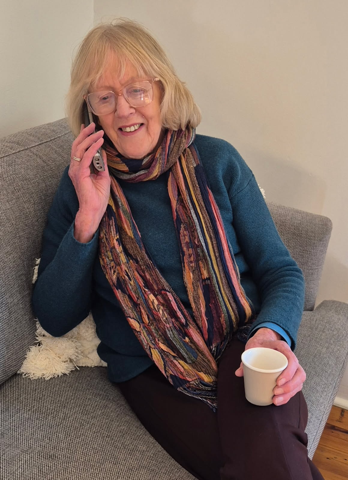 An older woman in a teal jumper and a bright striped scarf sits on a sofa laughing, a phone held to her ear and a cup of tea in her other hand.